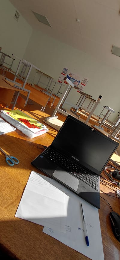 This is a medium shot, taken from a slightly elevated angle, of a classroom desk in Svetlogorsk, Russia. The desk is made of wood and is positioned in front of a row of identical student desks with metal frames and wooden surfaces. The classroom appears to be empty, and the desks are stacked in a way that suggests they are not in use. The lighting suggests it is daytime, with sunlight streaming in from the right side of the frame, casting long shadows across the desk.
On the desk in the foreground, there is an open black laptop with the brand name "AQUARIUS" visible on the lid. To the left of the laptop, a pair of blue scissors rests on a stack of papers and a colorful children's book. To the right of the laptop, a computer mouse is partially visible, connected by a black cable. In front of the laptop, a white sheet of paper is placed, with a blue ballpoint pen lying on it. Some handwritten notes are visible on this paper, as well as on another sheet of paper underneath. In the background, posters are displayed on the wall, and there are ventilation grilles and a smoke detector on the ceiling. The overall scene conveys a sense of an academic environment, possibly a classroom after school hours or during a break.