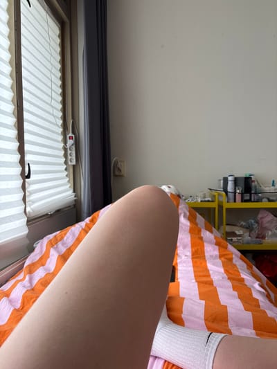 The image depicts a close-up view of a person's leg and arm resting on a bed with orange and pink striped bedding. The person is wearing a white sock. In the background, there is a window with white accordion blinds, a grey curtain, and an electrical outlet with a power strip. Further in the background, a yellow shelf unit holds various items, including bottles and containers. The scene appears to be indoors, possibly in a bedroom. There is no identifiable information to determine the location within the city of Enschede, Netherlands.