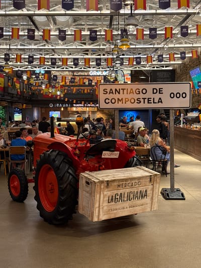 A red tractor is parked indoors in front of a wooden crate with "MERCADO LAGALICIANA SERVICIO A DOMICILIO" written on it. Above the tractor, a white sign with black text reads "SANTIAGO DE COMPOSTELA 000". Several people are seated at tables in the background, and flags hang from the ceiling. The environment appears to be a restaurant or market.