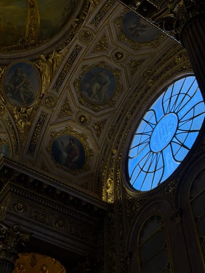 The image captures an ornate interior, likely a historic building or a museum, with a focus on the richly decorated ceiling and a large, blue-tinted arched window. The ceiling is a complex mosaic of hexagonal panels adorned with intricate gold detailing, possibly gilded plasterwork or stucco. Several circular paintings, depicting figures in classical attire, are embedded within the ceiling's design. These paintings are set within gold frames that echo the geometric patterns of the ceiling.
To the right of the frame, a large, elliptical window with a radial pattern of leaded glass dominates the upper portion. The glass is a vibrant blue, suggesting it is either stained glass or illuminated by a bright blue sky. Below this, a series of arched windows, also with geometric patterns in their panes, are visible.
The lower part of the image shows a dark, richly carved wooden element, possibly part of an entablature or a grand archway. This section features detailed reliefs, including what appear to be faces or masks and floral motifs, all rendered in dark wood with hints of gold. A large, fluted column, also dark and substantial, is visible on the far right edge of the image, adding to the sense of grandeur and architectural scale.
The lighting in the scene is dramatic, with strong contrasts between the illuminated window and the shadowed interior. This creates a sense of depth and highlights the textures and details of the ornamentation.
While the prompt mentions Mozzo, Italy, there are no discernible visual cues in the image that specifically place it in that location. The architectural style is reminiscent of European classical or baroque interiors, but without further context, a precise identification of the building or city is not possible from the image alone.