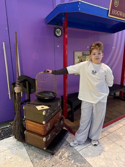 A medium shot captures a young boy, positioned at the center of the image, in a setting that seems to be a Harry Potter-themed display. The boy is wearing a long-sleeved white shirt with a small graphic on the front, paired with grey sweatpants and white sneakers. He is looking directly at the camera with a neutral expression.
The backdrop is a purple wall, and on the left side, there is a partial view of a structure that resembles a train platform with the word "Hogwarts" visible. Below the platform, there is a sign that reads "Platform 9 3/4."
In front of the boy, there is a display of Harry Potter-related items, including a birdcage, a wizard's hat, a stack of vintage suitcases, and a broom. The boy's hand is near the birdcage, as if he is interacting with the display.
The floor has a marbled pattern, and in the background, there are hints of other elements, such as benches or stands, that suggest this is a public space or exhibit.
There is no indication of the location being in the city of Родниковское сельское поселение, Ukraine, or any other specific location. The image is a scene that might be found in a themed exhibit or a Harry Potter event.