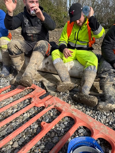 An outdoor scene in Baldurgan Hill, Ireland, depicts three adult males, likely laborers, taking a break while seated on light-colored cylindrical pipes. The central figure, wearing a black hooded sweatshirt and soiled black overalls, is drinking from a silver can, with his left hand raised in a gesture. To his right, another man, clad in a neon yellow long-sleeved top, an orange safety vest, neon yellow trousers, and a black baseball cap with a red bull logo, is holding a clear plastic water bottle to the side of his head. The leftmost man's torso and lower body are partially visible, wearing a blue top, light-colored work trousers, and blue boots. All visible work boots are heavily caked with mud. The foreground features an orange plastic temporary barrier segment lying on a ground composed of grey gravel and stones. A blue hard hat is visible in the lower right corner. The background consists of an earthen bank with sparse green vegetation under a clear sky.