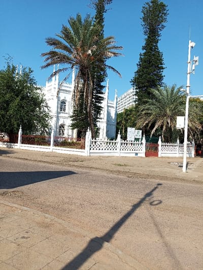 The image shows a white building with a white fence in front of it. The building has a unique architectural style with tall, pointed towers.  The building is located on a street in Maputo, Mozambique, at the coordinates -25.978, 32.586. There are palm trees and other trees in front of the building. The sky is blue and clear. The road is paved and there are shadows cast from the street lights. There are no people in the image.  The building appears to be a government building or a large house.  The environment is clean and well-maintained.