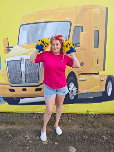 A woman stands in front of a large, yellow mural of a truck. She is wearing a bright pink t-shirt, denim shorts, and white slip-on shoes. Her hair is styled in braids and she has a red bandana tied around her head. She is holding two fluffy yellow and blue items, possibly dusters or cleaning tools, up near her face. The ground in front of the mural is a mix of dirt and small stones. The mural itself depicts the front and side of a yellow semi-truck with a prominent chrome grille. The background is a solid yellow, suggesting an outdoor setting, possibly a wall or a large banner. The specific location in Cheremushki, Russia cannot be determined from the visual information provided.