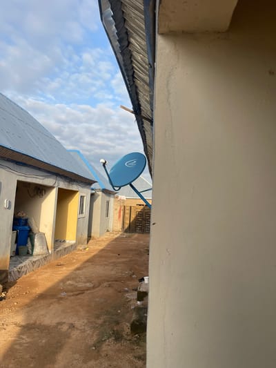 The image depicts an outdoor scene in Tavo, Nigeria, likely in the late afternoon as suggested by the long shadows cast on the sandy ground. The sky is partly cloudy with a mixture of white and grey clouds against a pale blue backdrop, indicating fair weather.

The foreground is dominated by the underside of a light-colored building, with a portion of a corrugated metal roof visible to the left. Below this, a blue satellite dish with the "dstv" logo is mounted on a pole, pointing upwards.

The midground reveals a series of small, attached structures, likely residential units, with light grey walls and some with yellow-painted openings. Windows are visible on some of these units. On the ground next to these structures, there are several items including plastic bottles, a blue plastic bin, and some white bags.

The background shows more of these structures and a brown, sandy ground with some vegetation and debris. The overall impression is of a residential area in a developing or rural setting. There is no discernible human activity in the image. The "dstv" logo on the satellite dish is the only visible text.