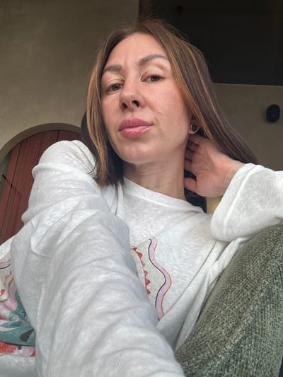 A medium shot portrait captures a woman with her head tilted, looking directly into the camera. She has medium-brown hair with lighter highlights, styled to fall over her shoulders. Her left hand is visible, resting behind her neck, and she wears a simple ring on her ring finger. Her lips are slightly parted, revealing a hint of a smile. Her left eye is in focus, displaying a warm brown hue.
She is dressed in a light-colored, possibly white or off-white, long-sleeved top made of a textured, somewhat sheer fabric. A colorful pattern, which appears to be part of a design on her top or another layer of clothing, is visible near her left shoulder and arm. To her right, a portion of a dark green, textured fabric, likely a couch or blanket, is visible.
The background is dimly lit, with dark walls or furnishings providing a stark contrast to her light top. A rounded object with vertical lines, possibly a piece of furniture or an architectural detail, is partially visible behind her right arm. The overall lighting suggests an indoor setting, possibly during the daytime with indirect light. No text is visible in the image.