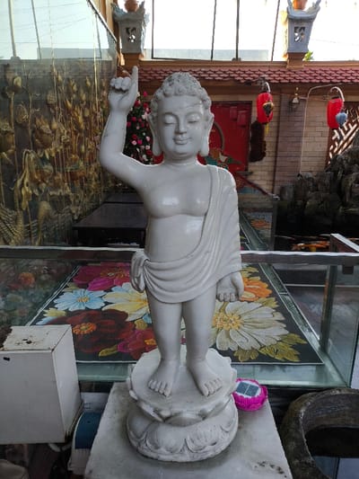 A white marble statue of a standing young Buddha, depicting Siddhartha Gautama with his right arm raised and index finger pointing skyward, stands on a two-tiered lotus pedestal. The statue is bare-chested with a draped garment across its left shoulder and waist, featuring a serene expression with closed eyes and tightly curled hair. The scene is set in Thành phố Quảng Ngãi, Vietnam.
In the immediate foreground, to the left of the statue's base, is a white rectangular box with rust stains. To the right of the base, a pink lotus-shaped solar-powered light is present, with a dark stone basin partially visible further right. Behind the statue, a clear glass barrier separates the foreground from the midground, revealing a vibrant floral-patterned surface beneath, likely the base of a pond or a decorative floor. Orange and white koi fish are visible in water behind the glass barrier to the right.
The background features a golden relief sculpture on the left, depicting lotus motifs and possibly a mythical creature. Two dark rectangular tables are positioned beneath this relief. The right background displays red architectural elements, including a wall or structure, adorned with hanging red lanterns and one blue lantern. Traditional roof tiles and decorative stone lanterns are visible above. A large bouquet of pink and red flowers is positioned centrally behind the statue's head. The upper background shows reflective glass panels, likely part of a building's upper structure.