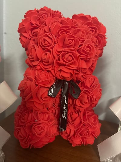 The image shows a close-up of a red rose teddy bear with a black ribbon tied around its neck that says "Just for you" in white lettering. The teddy bear is made of many small red rose-shaped petals and is sitting on a wooden surface. The background is a plain, light-colored wall. The presence of this specific item, a rose teddy bear, does not provide enough information to determine its location in Małkowo, Poland.