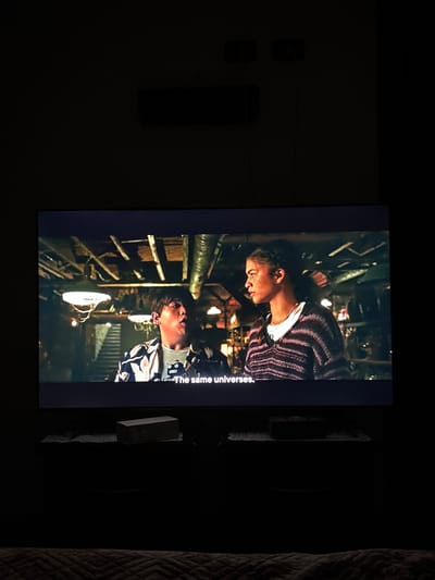 The image shows a television screen displaying a scene with two young individuals in what appears to be an enclosed, possibly subterranean, space with exposed pipes and metal structures. The scene is lit by what looks like industrial-style light fixtures. One individual, a young woman, is wearing a striped sweater with a white collar. The other, a young man, is wearing a patterned shirt. Subtitles are visible on the screen, reading "The same universe". The television is positioned on a dark surface next to what appears to be a textured blanket or cushion. In the background, on the wall to the left of the television, there are electrical sockets and a switch. The photograph was taken in an indoor environment. I have no information regarding the location in the city of Giugliano in Campania, Italy.
