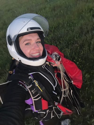 A woman, wearing a white helmet with a clear visor and a black and white skydiving suit, is lying on her back in a grassy field. A red parachute is partially visible on her right side. The woman is smiling and looking at the camera, holding her helmet with her left hand. The background is a blur of green grass, suggesting an outdoor environment, possibly a field outside of Limbaži, Latvia.