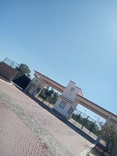 The image displays an entrance gate structure in La Venta, Mexico, under a clear, bright blue sky. The gate consists of a white-painted building featuring a reddish-brown tiled roof. A central archway, with a crest or seal visible at its apex, is flanked by black metal fencing sections. To the right of the archway, a small guard booth or office, complete with a rectangular window and a doorway, is integrated into the structure. The ground leading up to the gate is paved with reddish-brown interlocking pavers, exhibiting a darker, possibly wet or stained, linear strip down the center of the pathway. Green foliage, including trees and bushes, is visible behind the fences. No individuals or vehicles are present in the scene.
