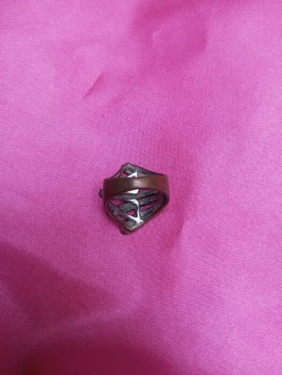 A single metallic ring is depicted resting on a fuchsia-pink fabric surface. The ring features a broad, reddish-brown metallic band. Attached to this band is a decorative, openwork element made of a lighter, silver-toned metal. This decorative component is approximately diamond-shaped, characterized by a geometric, lattice-like pattern of cutouts. The ring's metallic surfaces exhibit visible wear, scratches, and areas of discoloration consistent with tarnish. The fabric background shows slight creases and variations in texture. No other objects or individuals are present. The location is Qarabucaq qəsəbə, Azerbaijan.