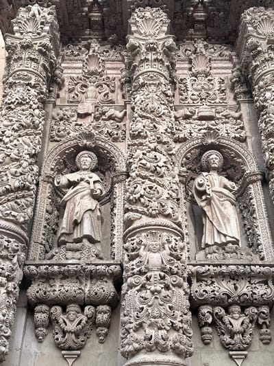The image shows a detailed view of the highly ornate facade of the Zacatecas Cathedral in Zacatecas City, Mexico. The facade is composed of intricate stone carvings, featuring columns, statues, and decorative motifs. Two prominent statues of male figures, likely saints, are visible within arched niches. They are dressed in robes and hold symbolic objects, though their identities are not clearly discernible from this angle. The stonework is heavily detailed, with layers of relief carvings depicting cherubs, floral elements, and geometric patterns. Fine mesh netting is visible in front of the statues and some of the decorative elements, likely to protect them from the elements or birds. The overall impression is one of elaborate Baroque architecture, characterized by its richness and complexity.