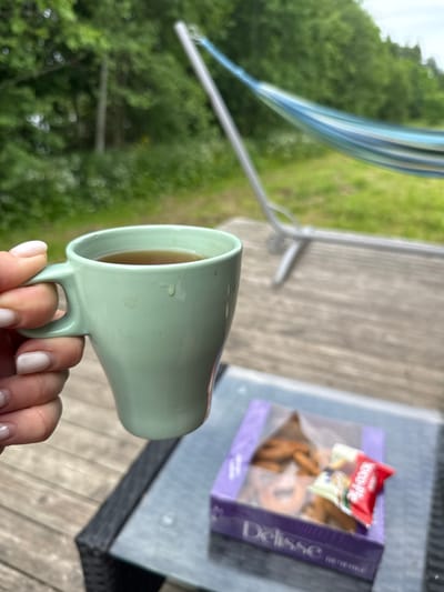 An overhead shot shows a hand holding a light green mug filled with a brown liquid, likely tea or coffee. The mug has a slight sheen and a small handle. In the background, a portion of a glass-topped table is visible, with a purple box of cookies on it. One cookie is partially unwrapped from a red and white wrapper. Beyond the table, a wooden deck leads to a green, grassy area with trees and a blue and white striped hammock. The overall setting appears to be outdoors, suggesting a leisurely moment. The image does not provide specific details about the city of Zamosh'e (Zaklinskoe S/P), Russia, beyond its potential association with such a scene.