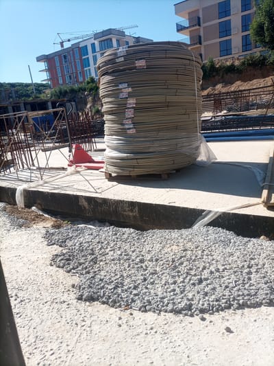 The image depicts a construction site under a clear blue sky. In the foreground, a large pile of coiled rebar, wrapped in clear plastic, dominates the scene. The rebar is stacked on a wooden pallet, with a red pallet jack visible to its left. Adjacent to the rebar, a concrete foundation runs horizontally across the frame, partially covered with light-colored plastic sheeting. A trench filled with gravel and some dirt runs along the front edge of the foundation. In the background, two multi-story buildings are under construction. One building, closer to the viewer, has a red brick facade and multiple windows. Another building, further back, has a light-colored facade with balconies and windows. Construction cranes are visible above the buildings. The overall environment suggests an active construction zone.