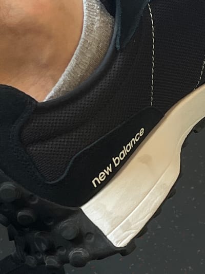A close-up, low-angle shot shows the side of a black New Balance sneaker with a white sole. The shoe is worn by a person, whose skin and grey sock are visible near the ankle opening. The sneaker features white stitching along the side panel and the "new balance" logo printed in white on a black tab. The sole has some dirt smudges. The background is dark and out of focus. The image was taken in Vejprnice, Czechia.