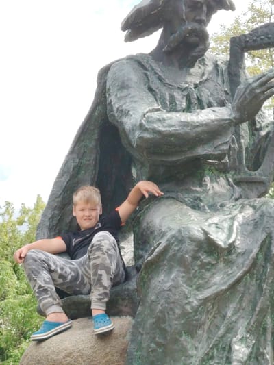 A young boy with blond hair is sitting on a rock next to a large bronze statue in Balyko-Shchuchynka, Ukraine. The boy is wearing a black t-shirt with an orange trim on the sleeves and camouflage pants. He has blue clogs on his feet. The statue depicts a man with a beard and a cap, holding a harp. The statue is weathered and has green patina in places. The background shows trees and a cloudy sky. The boy is looking at the camera and smiling.