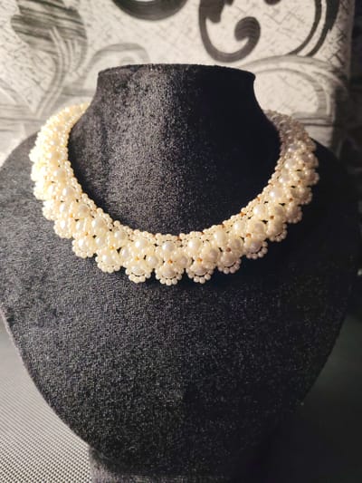 A close-up shot shows a pearl necklace displayed on a black mannequin bust. The necklace is intricate, composed of layers of cream-colored pearls of varying sizes, interspersed with tiny gold beads that create a floral pattern. The background is a patterned wallpaper in shades of grey and black, and the mannequin bust is placed on a textured grey surface. The lighting is dim, creating shadows and highlighting the texture of the pearls and the fabric of the mannequin. The location is not specified as Сокол, Russia, or any other city.