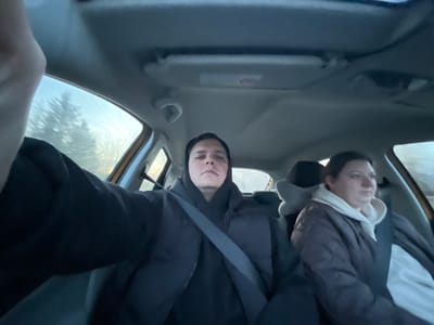 The image depicts the interior of a car, with the photographer positioned upside-down relative to the car's orientation. Two people are visible: one wearing a dark jacket and a seatbelt, and another to their left wearing a lighter jacket with a hooded sweatshirt underneath. The car's sun visor is visible in the foreground. Outside the car windows, blurry shapes suggestive of trees or foliage can be seen. Location in Yasenevo, Russia cannot be confirmed from this image.
