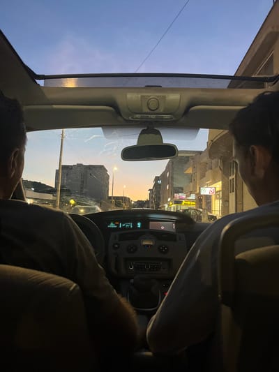 The image captures the interior of a car at dusk, looking forward through the windshield and sunroof. Two individuals are seated in the front of the car, their backs to the camera. The driver's side occupant has dark hair and is wearing a gray shirt. The passenger side occupant has dark hair and is wearing a blue shirt. The car's dashboard is visible, displaying a digital screen with text and icons, including "AUTO 22" and "EMISSIONS". The sunroof is open, revealing a twilight sky with hues of blue and orange. Outside the car, a street scene unfolds with buildings lining the right side of the road, some illuminated by yellow streetlights. A large, dark building is prominent on the left side of the street. The overall impression is of a car journey through a city during the transition from day to night. The location is identified as Pistula, Montenegro.