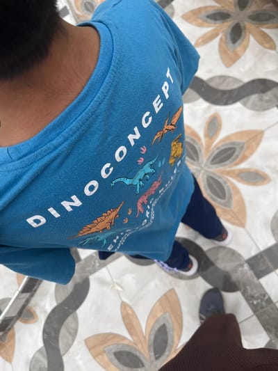 A person wearing a blue t-shirt with "DINOCONCEPT" printed vertically on it stands on a tiled floor. The tiles have a floral pattern with gray and beige accents. The person is also wearing dark blue pants and white-and-blue sneakers. Part of another person wearing a brown sweater and blue shoes is visible on the right side. The picture was taken in Quilmaná, Peru.