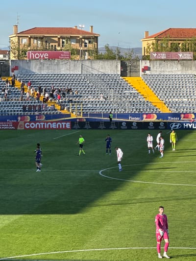 The image captures a soccer game in a stadium in Chaves, Portugal, showcasing several distinct sections.
In the foreground, on the right, a soccer field dominates the scene, populated by soccer players in action. The field shows some shadow.
On the left, stadium seating stretches into the distance, filled with spectators. Buildings are visible, one with a "hove" banner, while another displays "tintas Europa." A large "CONTINENTE" banner and other banner ads like "Tintas" and "HYLI" adorn the stadium infrastructure.
