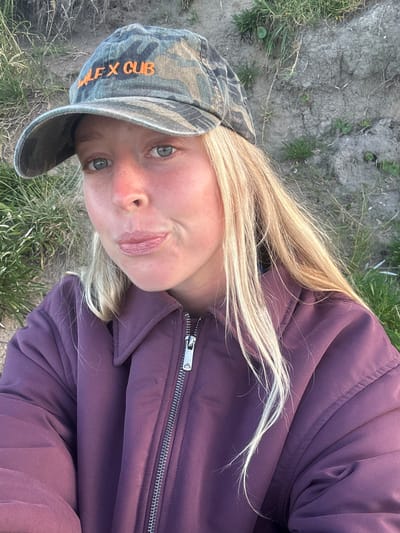 A blonde-haired individual wearing a camouflage baseball cap with orange lettering "FLEX CUB" and a purple jacket is positioned outdoors on a grassy area with patches of dirt. The location is unidentifiable within Wollongong City Council, Australia, from the given image alone. The individual is looking directly at the camera with a slight pout.