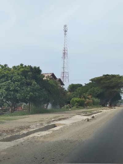 The image depicts an outdoor scene in Cotpuuk, Indonesia, on a hazy or overcast day. The primary subjects are a tall, lattice-style telecommunications tower in the background, and a roadside with ongoing construction or maintenance work. Lush green trees are prominent, lining the roadside and obscuring some of the buildings behind them.
The telecommunications tower, with its white and red lattice structure, stands out against the pale sky. To its left, partially visible, are the rooftops of houses with reddish-brown tiles and gabled roofs.
The roadside itself appears to be under construction or repair. A concrete drainage channel or gutter is being built or cleaned alongside a dirt and gravel path. Several large, cylindrical concrete objects, possibly for construction, are placed along the edge of the road. Further down the road, there are hints of small structures and more vegetation, suggesting a somewhat rural or semi-urban environment.
The overall mood is neutral, conveying a sense of everyday activity and infrastructure development. There is no visible text in the image. The time of day appears to be daytime, but the overcast sky obscures direct sunlight, creating a diffused light.