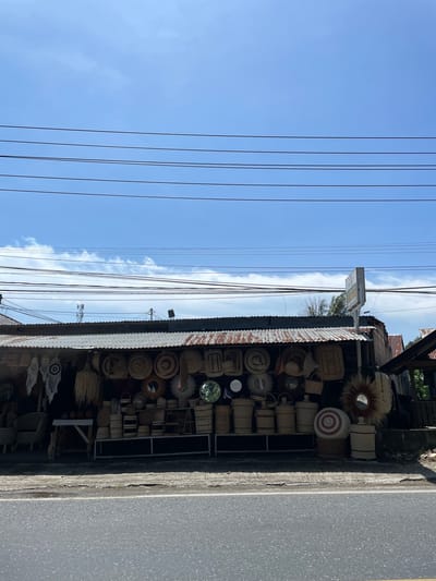The scene captures an outdoor marketplace in Tanjong, Indonesia, featuring a shopfront laden with woven goods and mirrors. The display extends under a corrugated metal roof, with a gray asphalt road running adjacent to the shop. Above the structure, electrical wires stretch across a blue sky scattered with white clouds.