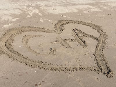 In Rezovo, Bulgaria, a heart shape is drawn in the sand on a beach. Inside the heart, the letters "G" and "A" are also drawn. The sand is light brown and textured, with some small shells and pebbles scattered on the surface. The sunlight creates shadows, indicating a bright, sunny day. The area surrounding the heart appears to be undisturbed sand, with some faint footprints in the distance.