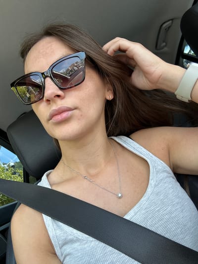 A woman wearing sunglasses and a grey tank top is inside a car. She is wearing a necklace and a smartwatch. A seatbelt is visible across her chest. The interior of the car is dark leather, and through the window, greenery and houses are visible. The scene is set in Stanišići, Montenegro.