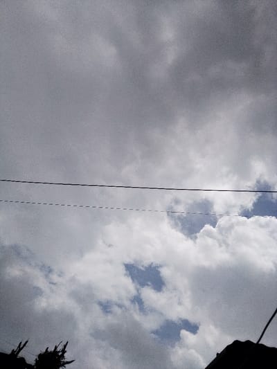 The scene depicts an upward-angled view of an overcast sky in Nachu ward, Kenya. The upper two-thirds of the frame are dominated by dense, grey cloud formations. Several irregular patches of clear blue sky are visible in the lower half of the frame, interspersed among the clouds. Two utility lines extend horizontally across the mid-ground: a solid black line positioned above a dashed grey and black line. The upper portion of dark, silhouetted foliage is visible at the bottom-left corner of the frame. A dark, silhouetted section of a building roof or structure is visible at the bottom-right corner. No individuals or active subjects are present. The image exhibits visible digital noise.
