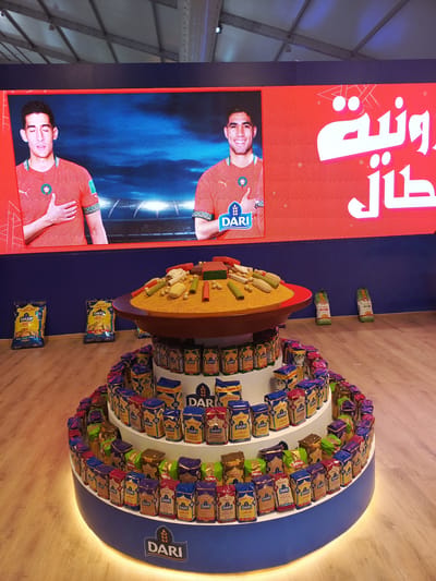 This media file captures an indoor promotional display, likely at an event or exhibition in Al Machouar – Stinia, Morocco. In the background, a large LED screen features two men, presumably Moroccan soccer players, wearing red national team jerseys with the Moroccan crest. The man on the right is smiling and giving a thumbs-up, while the man on the left has his eyes closed and a hand over his heart. The screen also displays the "DARI" brand logo and partial Arabic text, which appears to be part of a phrase like "وطنية" (national) and "طال" (long).
In the foreground, a prominent, multi-tiered circular display stand, illuminated from below, showcases numerous small packages of various couscous and semolina products from the "DARI" and "NEMA" brands. The top tier is adorned with a large, shallow dish filled with golden couscous-like grains, decorated with colorful cylindrical and block-shaped elements resembling vegetables or sweets. On the wooden floor, several large bags of "DARI" brand couscous are also visible. The overall setup suggests a product launch or promotional event for "DARI" couscous, possibly leveraging national pride or sports associations. The ceiling structure in the background indicates a temporary event space, like an exhibition hall or tent.