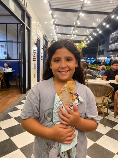 The image shows a young girl holding a lizard wrapped in a light blue towel. She is standing in an outdoor dining area, possibly a restaurant or cafe, in Punta Hermosa, Peru. The area is characterized by white tile walls with blue accents. There are strings of lights strung across the ceiling, creating a warm ambiance. Other people can be seen in the background, including a man sitting at a table and a woman standing. The girl is smiling, and the lizard appears to be docile. The floor is tiled in a black and white checkered pattern.