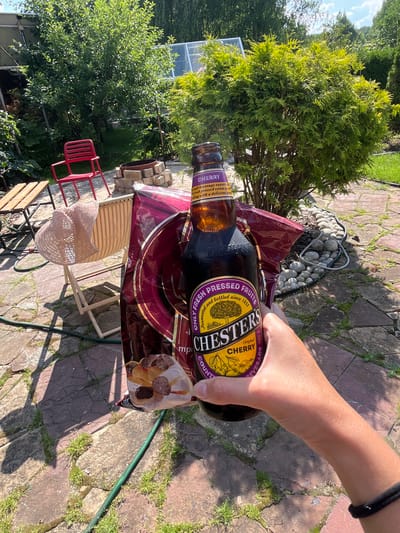 In the city of Вельяминово, Russia, a person's hand holds a bottle of Chesters Cherry beverage and a partially open bag of chocolates outdoors. The scene is set in a backyard with a paved patio, scattered stones, and green grass. Sunlight casts shadows from trees and other objects. In the background, there is a red chair, a wooden table with chairs, and a greenhouse. The overall impression is one of a casual, sunny day.