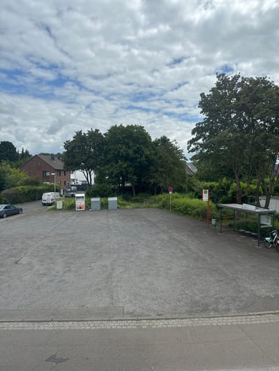 A cloudy sky with scattered white clouds is visible above a street scene in Leverkusen, Germany. In the foreground, a paved parking lot occupies the majority of the image. To the left of the parking lot, a grey car is parked. Further back, several buildings are visible, including a brick house with a white facade and a modern white building. A white van is parked in front of the brick house.
In the middle ground, three large, grey trash receptacles are lined up. To the right of the trash receptacles, a bus stop shelter with a metal frame and a translucent roof is present. Three benches are visible inside the shelter. Two bicycles are parked next to the bus stop. A red pole with a circular sign featuring a red border and a red diagonal line through a red circle is situated to the left of the bus stop. A bus stop sign with the letter "H" in a yellow circle is also present.
The background is dominated by lush green trees and vegetation. The ground is paved with asphalt and bordered by cobblestones and a grey sidewalk.