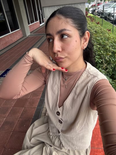 A woman with dark hair pulled back, wearing a light beige dress or jumpsuit with button details, is positioned at an angle in the image. She has on a light brown undershirt. She's adorned with silver rings and a layered necklace. The background shows a brick wall, windows, and greenery, indicating an outdoor or urban setting. The overall aesthetic is casual and fashionable.
