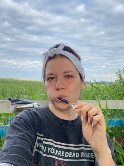 A selfie-style medium shot shows a woman holding a smoking pipe to her mouth, set against a backdrop of tall green grass and a cloudy sky. The woman, who appears to be outdoors, is wearing a dark t-shirt with a graphic print. A white bandana with a black pattern is tied around her head. She has a nose ring and hoop earrings. In the background, there is a white fence and some white objects, possibly bags or containers. The overall lighting suggests it is daytime, likely overcast. No specific location in Абзаново, Russia can be determined from the image.