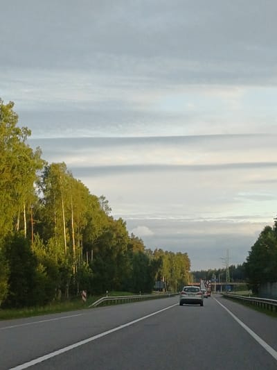 The image shows a view from a moving vehicle on a highway in Saulkrasti, Latvia. The highway has two lanes in each direction, separated by a median. There are guardrails on both sides of the road. On the left side of the road, there is a dense forest of birch and coniferous trees. On the right side, there are also trees and some buildings visible in the distance. The sky is overcast with a few patches of blue sky. There are two cars visible on the highway in front of the viewer's vehicle. The car directly in front is a silver SUV, and behind it, another vehicle, possibly a truck, is visible. The overall lighting suggests it is daytime, likely late afternoon due to the golden hue on the trees. There are no people visible outside of the vehicles.