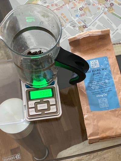The image shows a glass measuring jug on a digital kitchen scale next to a package of Kyra Haja Nai Xiang Wu Long tea. The scale reads "0.00" and a portion of a map can be seen under the jug and tea, with the street name "Matije Korvina" visible, placing the location in the city of Subotica, Serbia. A partial view of a white object or surface is visible in the lower right corner of the image.