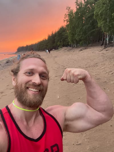 In Luzhki, Russia, a man with a beard and blond hair stands on a sandy beach at sunset. He is wearing a red tank top and a neon green necklace. He is flexing his bicep for the camera. In the background, there are trees and a body of water. The sky is a vibrant orange and pink. Several other people are visible in the distance, walking along the beach.