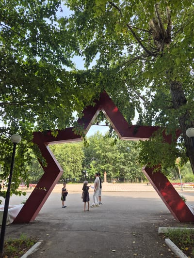 The image shows a large, red, five-pointed star monument in an outdoor setting. The star is a three-dimensional structure, appearing to be made of metal, with thick beams forming its points. It is positioned at an angle, suggesting it is an archway or entrance. Lush green trees with dense foliage surround and partially obscure the top of the star. The sky is visible through the leaves, appearing bright and possibly overcast.
In the foreground, an asphalt pathway leads towards the star. To the left, a black lamppost with a white spherical light fixture is visible, partially covered by leaves. To the right of the star, a large tree trunk with textured bark is prominent.
Several people are present in the scene, standing in front of and near the star. On the left side, a young boy in a dark t-shirt and shorts stands facing the star. Further back, a group of three people, two women and one man, are positioned. One woman is wearing a dark dress and a headscarf, another woman is in a light-colored dress, and the man is wearing a light t-shirt and pants. They appear to be interacting or observing the monument.
The background shows a paved area, likely a park or public square, with more trees and some flowerbeds visible in the distance. The overall environment suggests a sunny day in a green, urban or park-like area. The monument appears to be a significant landmark in the location. It is a monument in the city of Novoishimskoye, Kazakhstan.