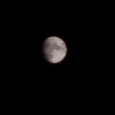 A photograph of the moon against a black sky. The moon is partially obscured by clouds, giving it a mottled appearance. A faint red glow encircles the moon, suggesting the presence of an atmospheric phenomenon or a photographic effect. The image is dark and still, with the moon being the sole discernible subject. The composition is simple, focusing attention solely on the celestial body. There is no visible text in the image.