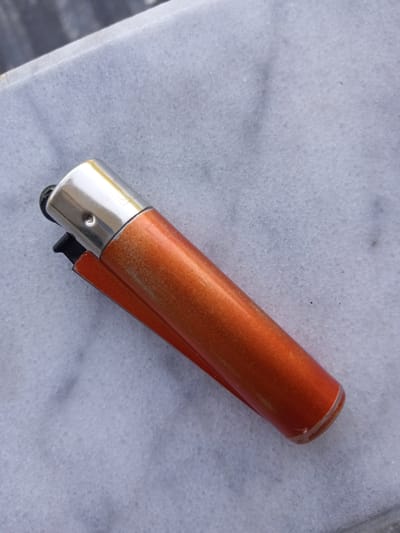 A single orange cigarette lighter with a silver top rests on a textured grey surface, likely marble or granite. The lighter is positioned diagonally, with its top left corner slightly raised. The surface beneath it appears speckled and slightly uneven. No people are present in the image. The setting is indoors, on a countertop or table.
