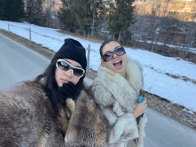 The image depicts two females posing outdoors on a snowy hillside in Saanen, Switzerland. The female on the left has long dark hair, wears a black knitted beanie, white-framed sunglasses with dark lenses, black gloves, and a multi-toned brown fur coat. Her right gloved hand rests near her chin, and her lips are slightly parted. The female on the right has dark hair pulled back, wears large dark-framed sunglasses, and a light-colored fur coat with a prominent collar. Her mouth is wide open in an expressive manner, and she holds a mobile phone in a blue-green case in her left hand. Both individuals are facing the camera. They are positioned next to a grey paved road surface. The immediate ground around them consists of patchy snow cover and areas of exposed earth. Thin wooden posts are visible along the edge of a snow-covered slope to the left. The background features snow-dusted forested mountains populated with both coniferous and bare deciduous trees. Several traditional wooden buildings with dark roofs are discernible nestled among the distant trees on the slopes. The scene is illuminated by daylight.