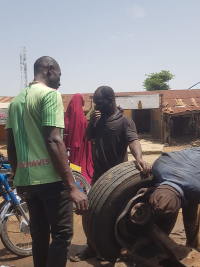 In Tukur Tukur, Nigeria, three men are engaged in activity outdoors. One man, wearing a green and white striped polo shirt with the inscription "MAHAVIS" on the back, stands facing away from the camera, looking towards the other two men. He is wearing dark pants and sandals. To his right, another man in a dark brown polo shirt is bent over, appearing to speak or gesture towards a large tire. He is also wearing dark pants. The third man, whose face is not visible, is wearing a blue shirt and is crouched down, working on the tire which is resting on metal machinery. The background shows a single-story building with a corrugated metal roof and a sign that reads "CAPENTRY & SERVICES NIG. LTD." A cell tower is visible in the distance against a clear sky. A blue motorcycle with a chrome fender is parked to the left of the frame. The ground is dusty and appears to be unpaved.