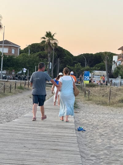 The image captures a scene on a beach boardwalk in Castelldefels, Spain, during the late afternoon or early evening. The sky is a soft, pale orange, indicative of sunset. A wooden boardwalk, composed of parallel planks, runs diagonally from the lower left to the upper right of the frame, bordered by sand on both sides.

In the foreground, a man in a grey t-shirt and dark shorts walks away from the camera on the boardwalk. Beside him and slightly ahead, a woman in a light blue, flowing dress walks in the same direction, carrying a woven straw bag. Behind them, a woman in a white hat and blue top is partially visible. Further down the boardwalk, several other people are walking and standing.

To the left of the boardwalk, there are sand dunes with sparse vegetation and a rope fence. In the background, a row of buildings, including a house with a red tile roof, are visible amongst lush green trees and palm trees. A street with parked motorcycles and a signpost with the number "29" and text in Spanish indicating "Playa de la Pineda" (Pineda Beach) are also present, confirming the location. The overall atmosphere suggests a leisurely summer evening by the sea.