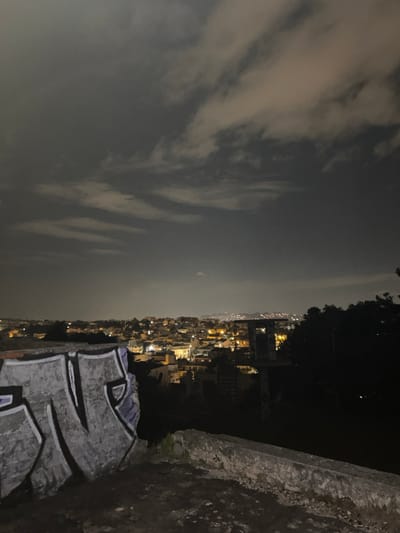 The image depicts a nocturnal panoramic view of the Gerakas Municipal Unit, Greece, from an elevated vantage point. In the immediate foreground, a weathered concrete structure, possibly a wall or ledge, exhibits extensive graffiti art, primarily in shades of gray, white, and purple. The surface appears rough and deteriorated. Beyond this structure, the midground presents a sprawling urban landscape illuminated by numerous artificial lights from buildings and streetlights, indicating a dense residential and commercial area. Silhouetted trees and dark architectural elements are visible amongst the illuminated city structures, particularly along the right side. The distant background features a faint horizon line with minimal illumination, suggesting elevated terrain or further urban sprawl. The sky, occupying the upper two-thirds of the frame, is dark and heavily overcast with multiple layers of clouds, some reflecting ambient light from below. A single, faint, pinpoint light source is discernable in the upper central portion of the sky. There are no individuals, active human figures, or discernible interactions within the scene. The environment is a static, dimly lit cityscape under a cloudy night sky.