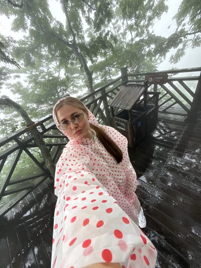 A medium-shot, slightly tilted photo shows a woman wearing glasses and a white raincoat with red polka dots, appearing to be in Yongding District, China. The scene appears to be on a wooden structure, possibly a raised walkway or bridge, with trees visible in the background and what appears to be fog. A small wooden cabin-like structure with a sign on top is nearby. The wooden floor of the structure is wet. The photo is taken from a high angle, looking down at the woman.