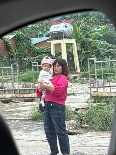 A woman in a pink shirt and blue jeans holds a baby wearing a pink hat and white outfit, standing outdoors. Behind them, a large metal water tank is supported by a yellow structure. The area appears somewhat unkempt, with debris and vegetation visible. The scene is framed by the interior of a vehicle, suggesting the photo was taken from inside a car. The location is identified as Tân Lập Commune, Vietnam, indicated by specific architectural and environmental cues typical of the region.